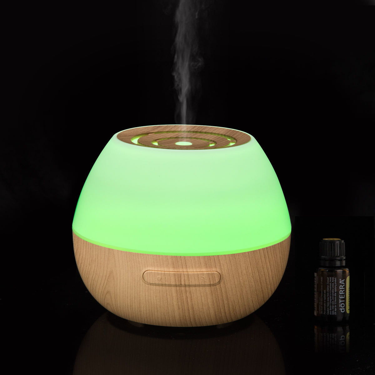 Diffuseur d'huiles essentielles à base effet bois, lumière verte et brume, à côté d'un flacon d'huile
